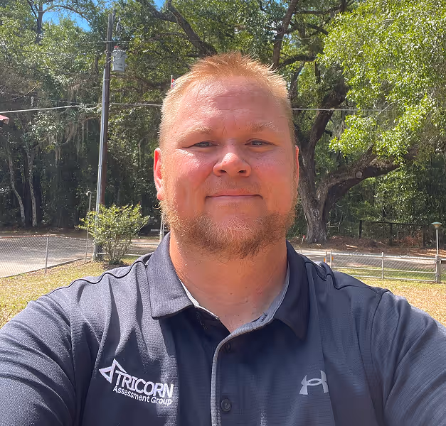 Man with short blond hair and beard wearing a Tricorn Assessment Group polo outside with trees behind.