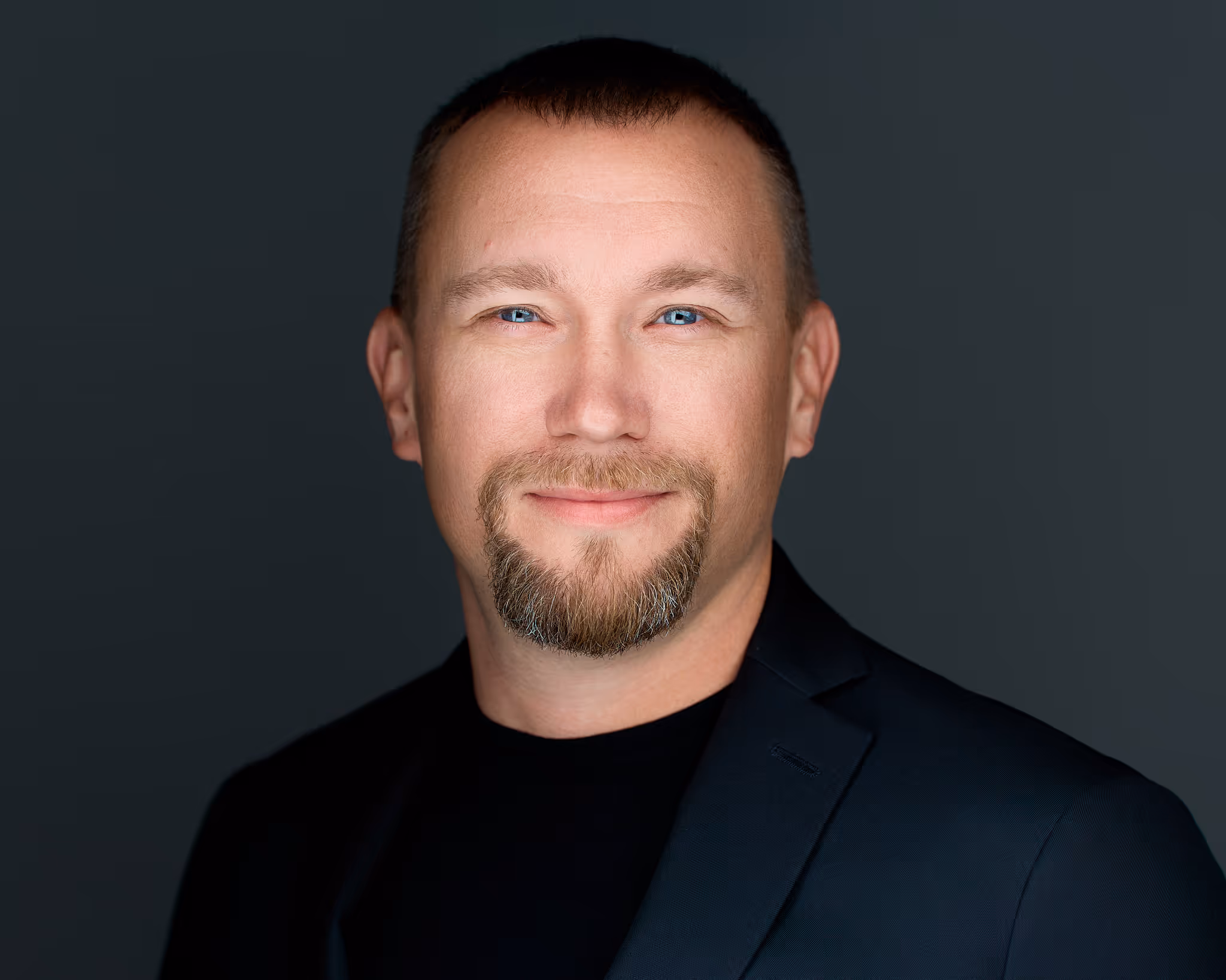 Smiling man with blue eyes, short brown hair, and goatee wearing a black jacket and shirt.