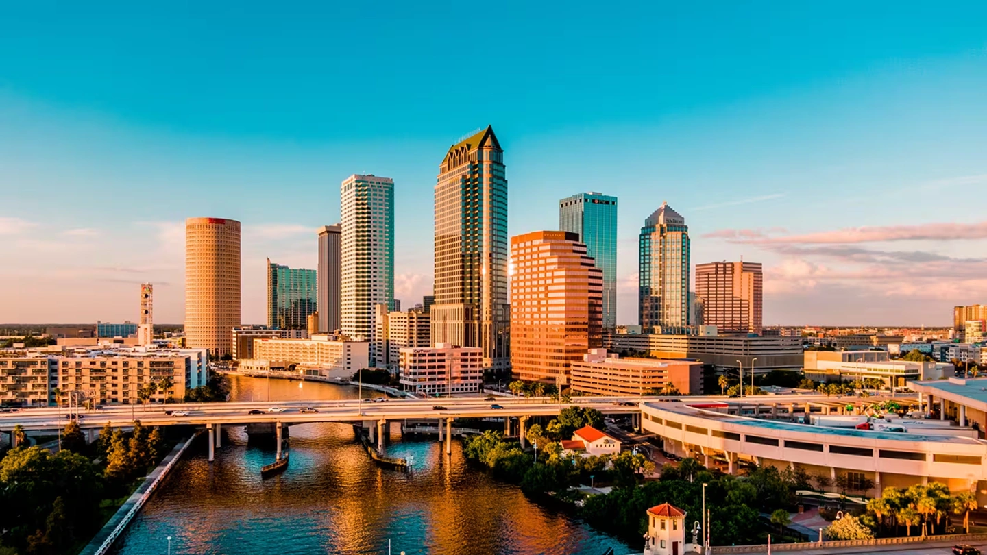 City skyline at sunset with a river and bridge in the foreground under a clear blue sky.