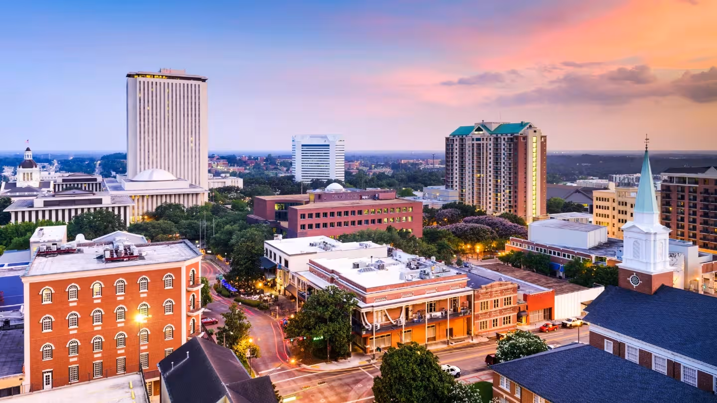 City skyline at sunset with a river and bridge in the foreground under a clear blue sky.
