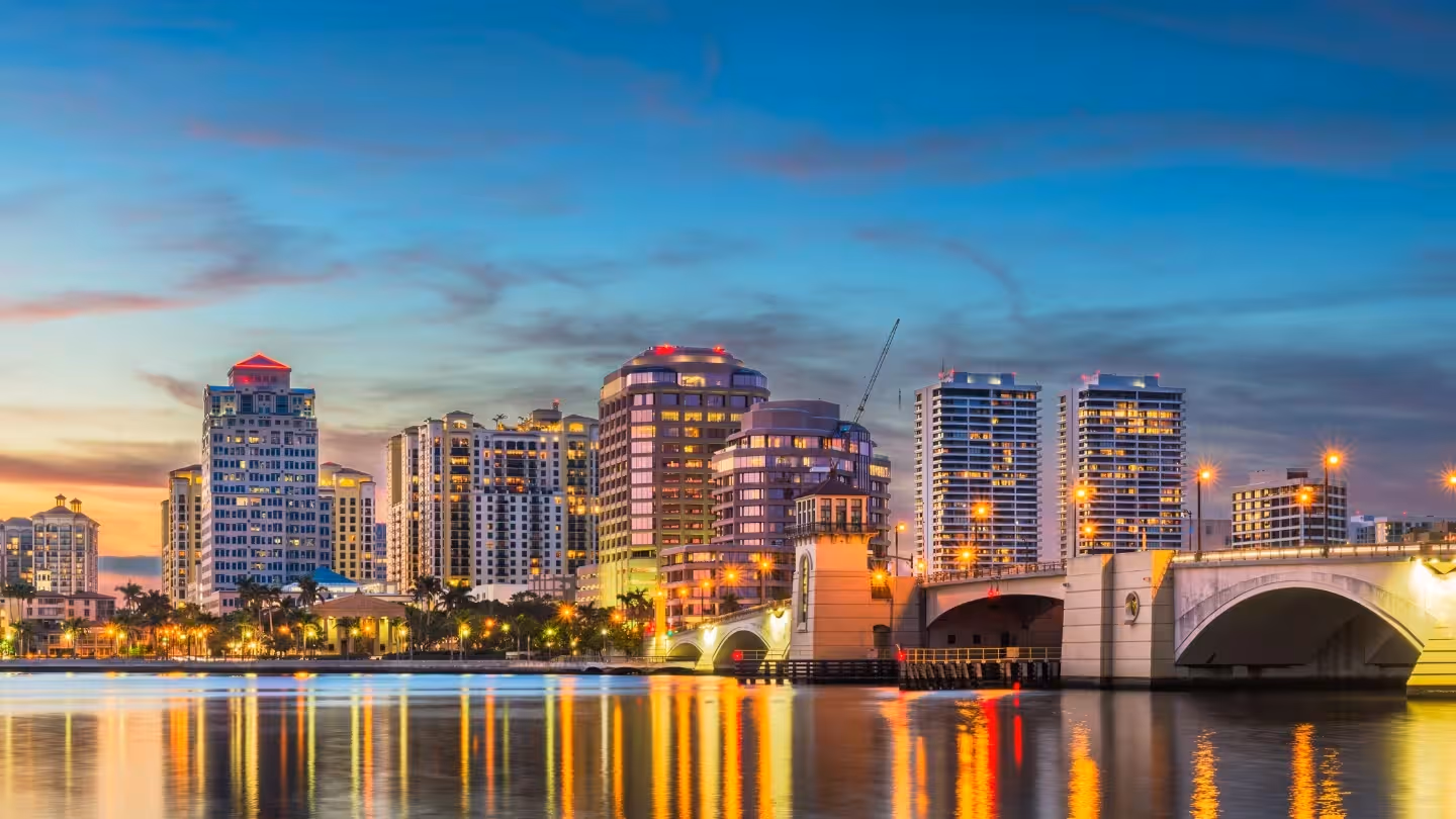 City skyline at sunset with a river and bridge in the foreground under a clear blue sky.