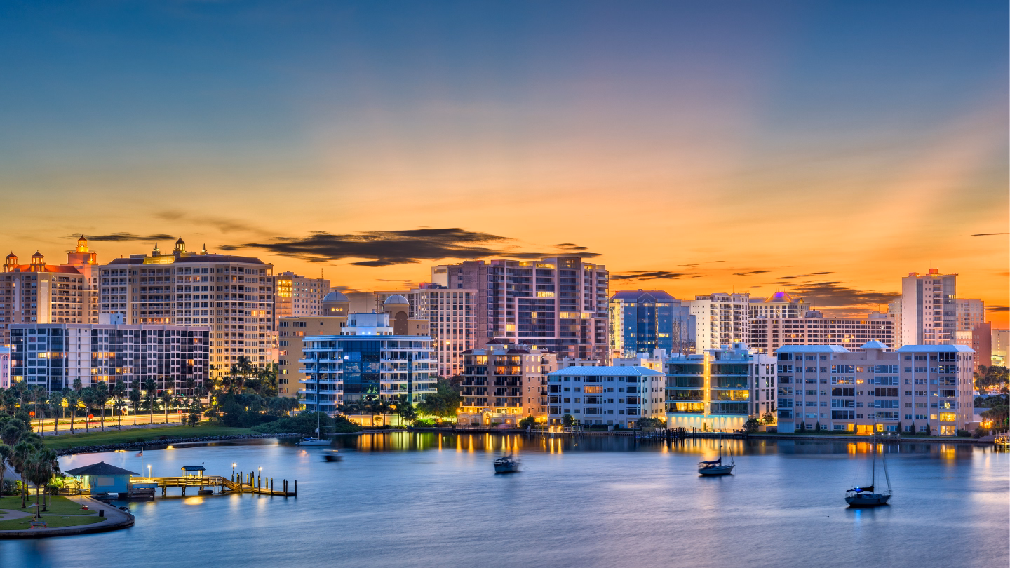 City skyline at sunset with a river and bridge in the foreground under a clear blue sky.