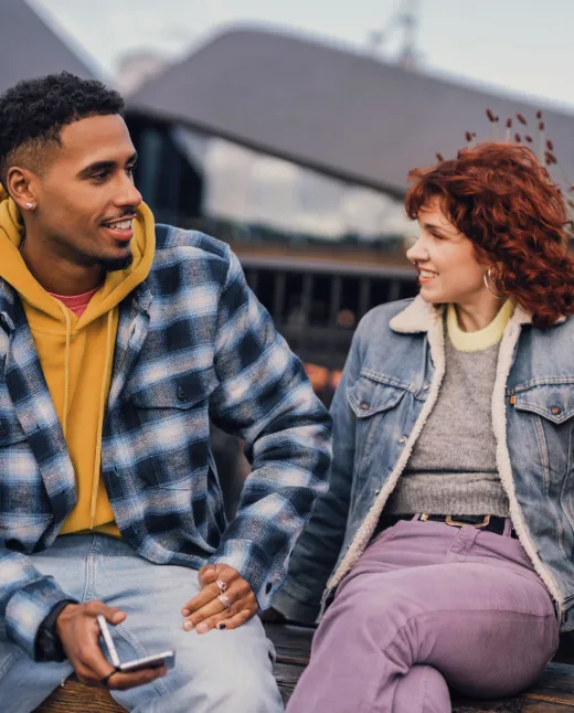 A man in a blue plaid jacket and mustard hoodie sitting and smiling while talking to a woman in a denim jacket and purple pants on a bench outdoors.