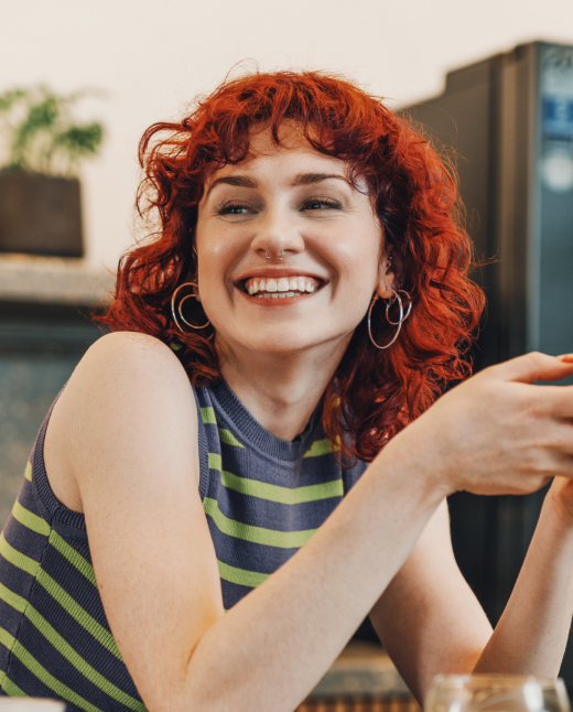 Smiling young woman with curly red hair wearing hoop earrings and a striped sleeveless top holding a cup.