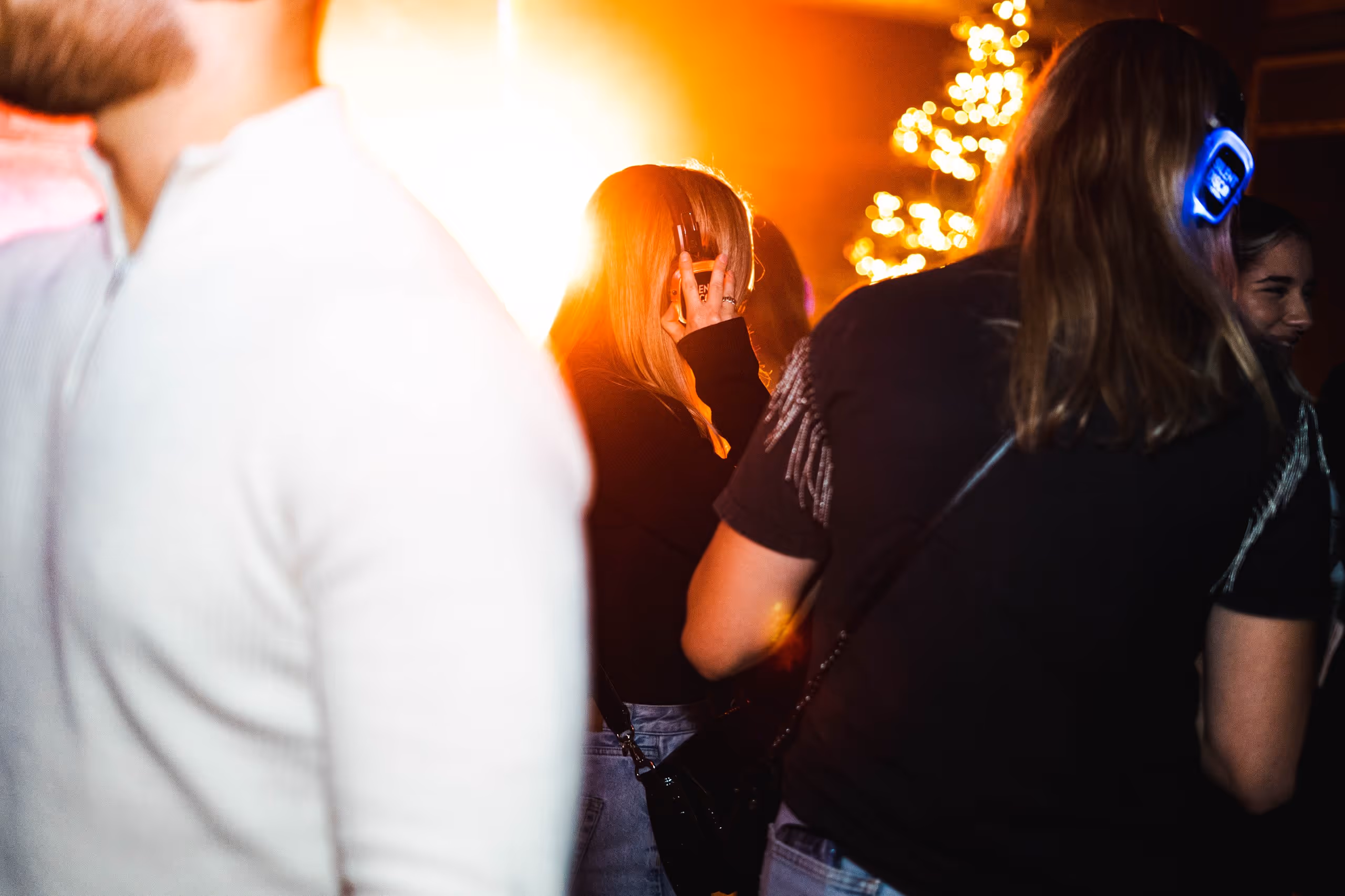 Jonge mensen dansen en socializen op een silent disco evenement, waarbij sommigen kleurrijke koptelefoons dragen tegen een levendig verlichte achtergrond met een feestelijke sfeer.