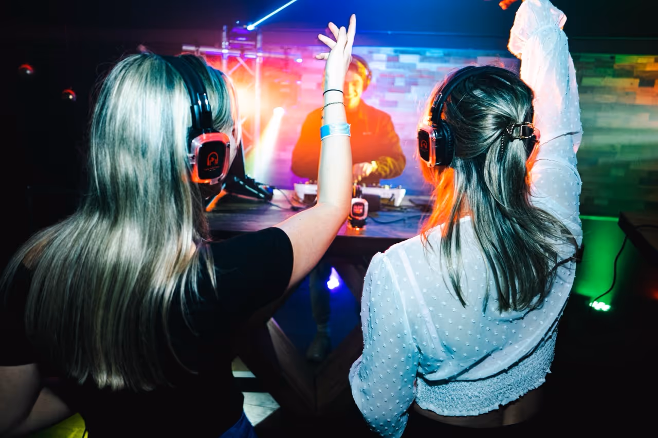 Twee vrouwen dansen met draadloze koptelefoons op een silent disco evenement, waarbij een DJ op de achtergrond muziek draait.