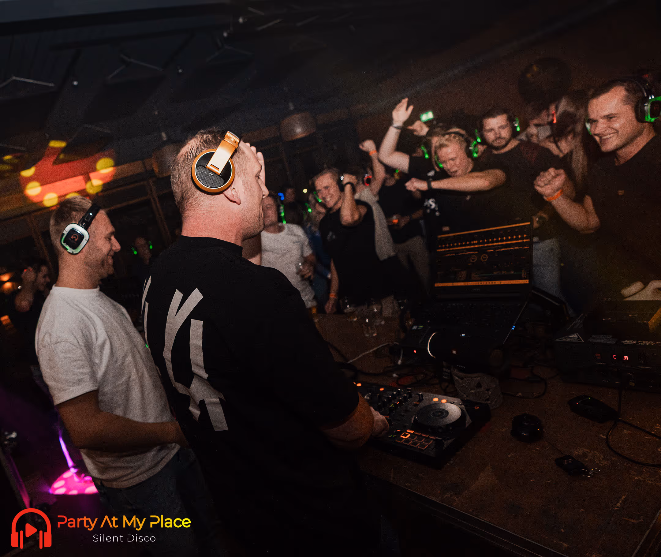 Op de afbeelding is een levendig silent disco evenement te zien waarbij deelnemers dansen en plezier hebben terwijl ze allemaal draadloze koptelefoons dragen; een DJ bedient de muziekapparatuur aan de voorzijde van de ruimte.