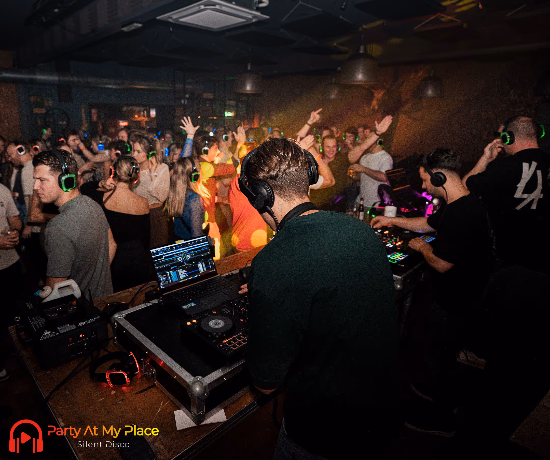 In de foto is een levendige silent disco te zien met meerdere mensen die dansen en verschillende kleuren koptelefoons dragen in een sfeervol verlichte nachtclub terwijl een DJ aan een mengpaneel werkt.