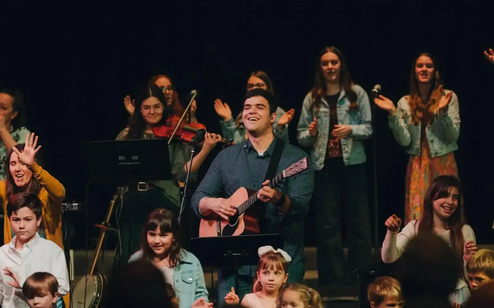 A joyful man is playing an acoustic guitar and singing on stage, surrounded by a diverse group of people, including children and adults, some clapping and others playing instruments like a violin. The background is dark, highlighting the performers who are engaged in a lively musical performance.