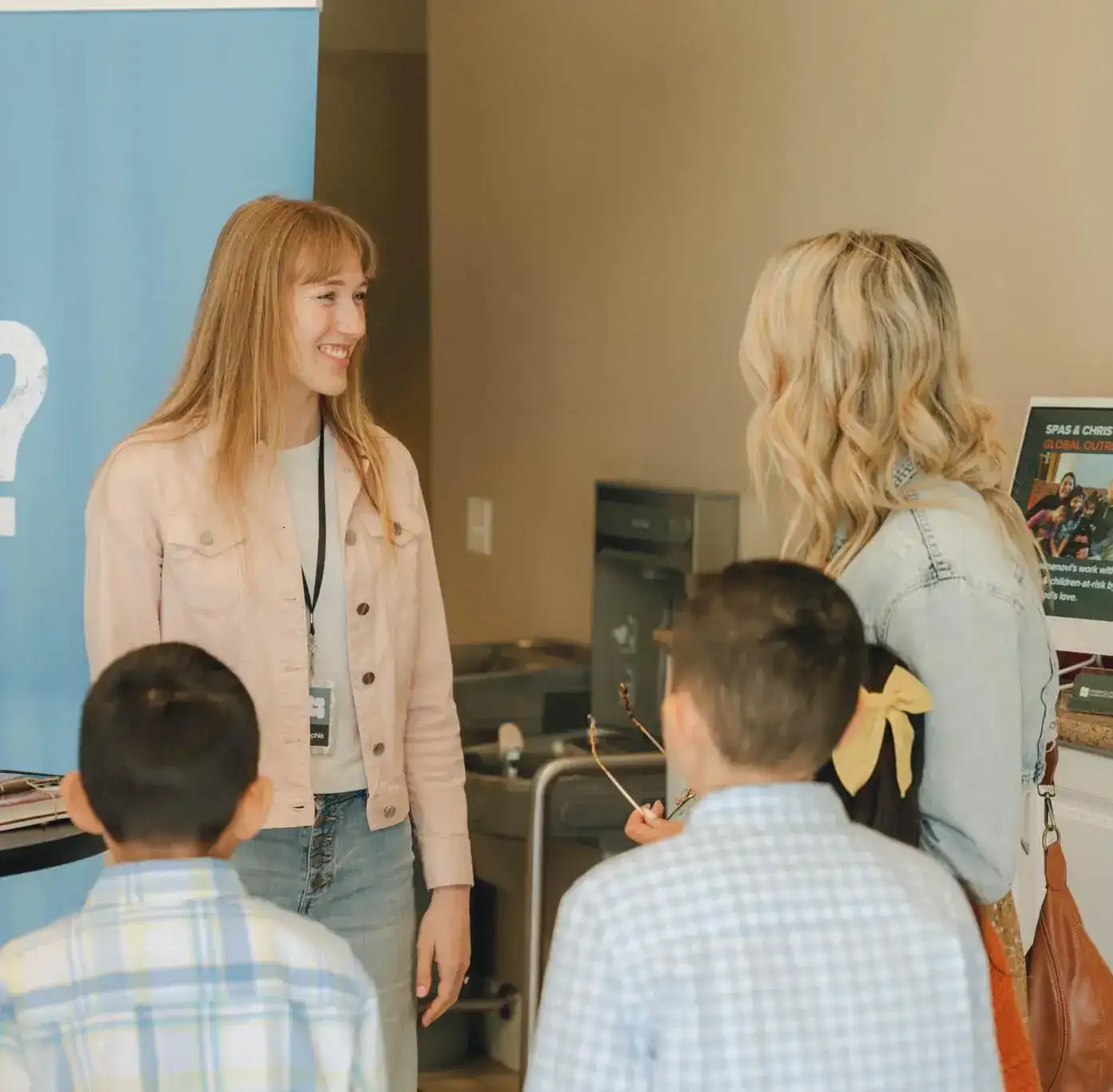 A smiling woman with a lanyard is talking to another woman and two young boys in a casual indoor setting, with a blue banner and informational poster in the background.