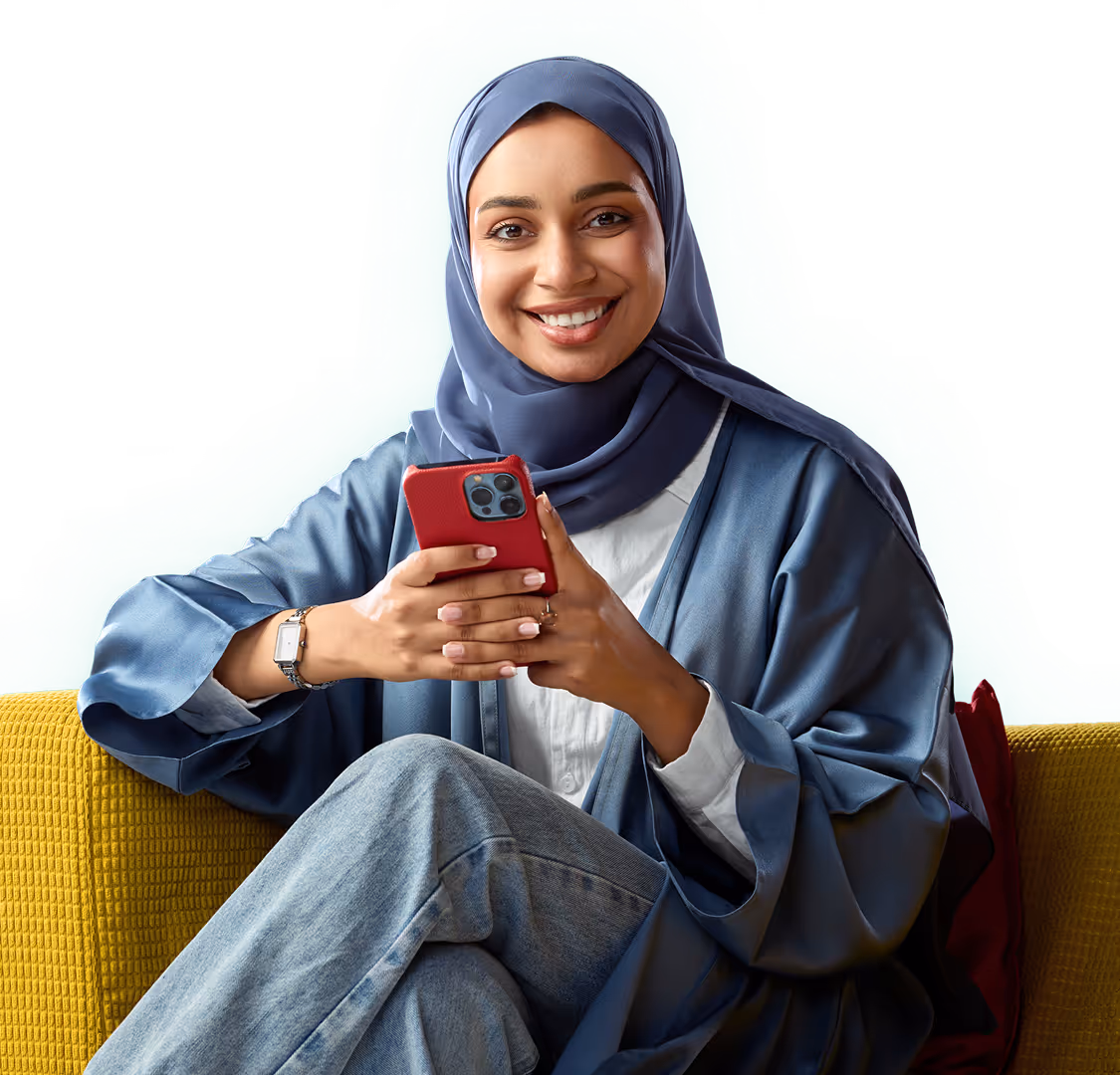 امرأة مبتسمة بحجاب وعباءة زرقاء تجلس على أريكة صفراء وتمسك هاتفاً أحمر.
