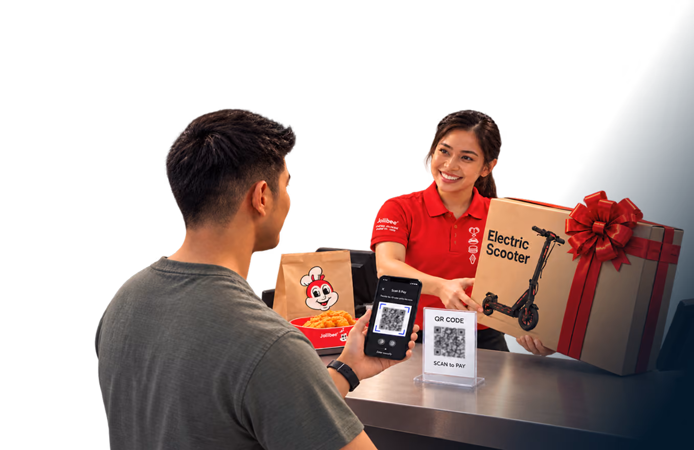 رجل يمسح رمز استجابة سريعة (QR code) بهاتفه عند منصة "جوليبي"، بينما تقف موظفة مبتسمة ترتدي زياً أحمر وتحمل صندوقاً مكتوباً عليه "سكوتر كهربائي" بجانب عبوات طعام "جوليبي" وصندوق هدايا كبير مزين بشريط أحمر.