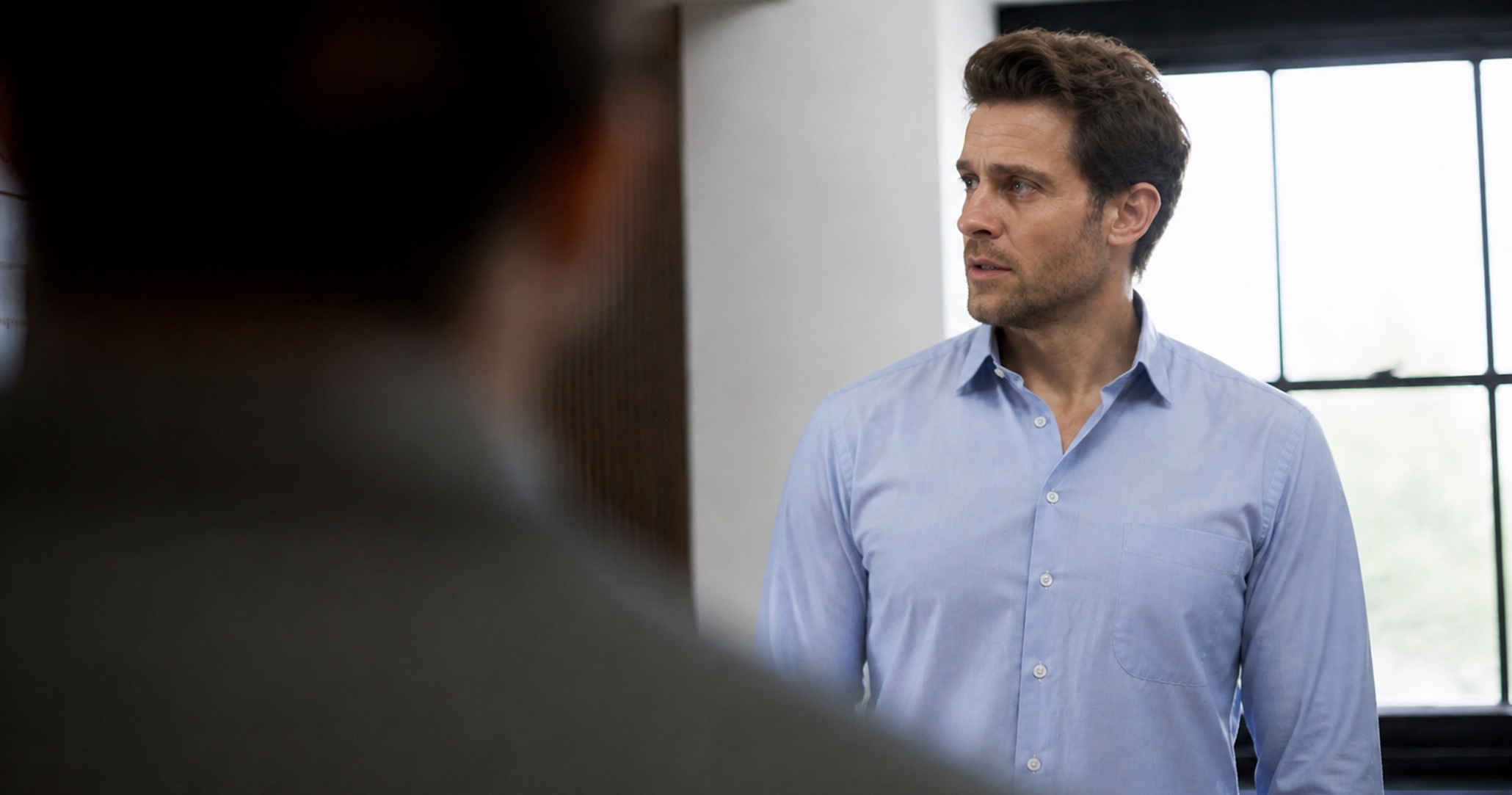 Man in blue shirt looking thoughtfully to his right near a window with grid panes.