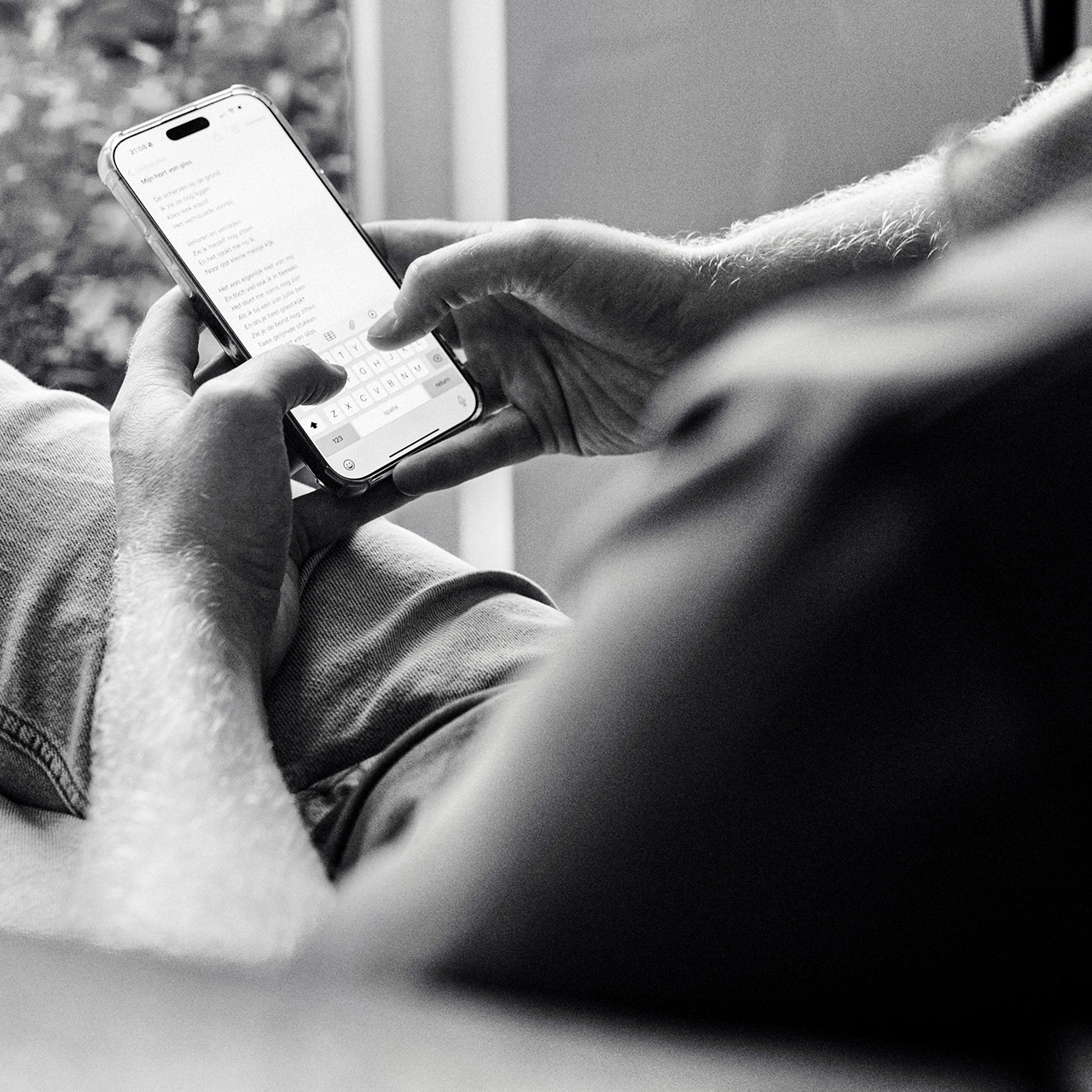 Zwart-witfoto van Adam Dekker die met beide handen een smartphone vasthoudt en een liedtekst typt, met tekst zichtbaar op het scherm.