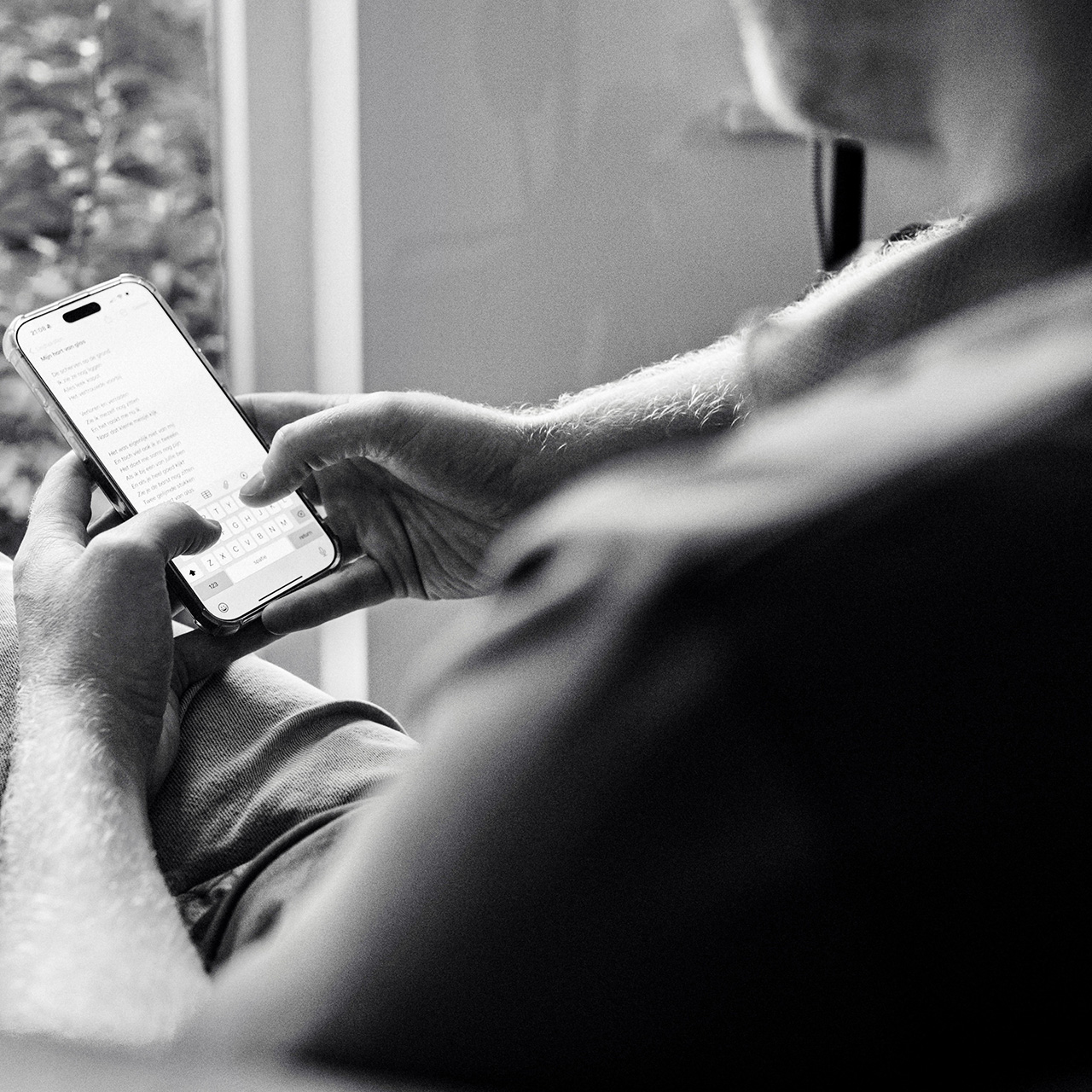 Zwart-witfoto van Adam Dekker die met beide handen een smartphone vasthoudt en een liedtekst typt, met tekst zichtbaar op het scherm.