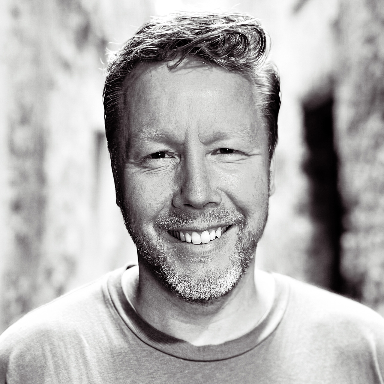 Portret van een glimlachende Adam Dekker met kort kapsel en baard, gefotografeerd in zwart-wit.