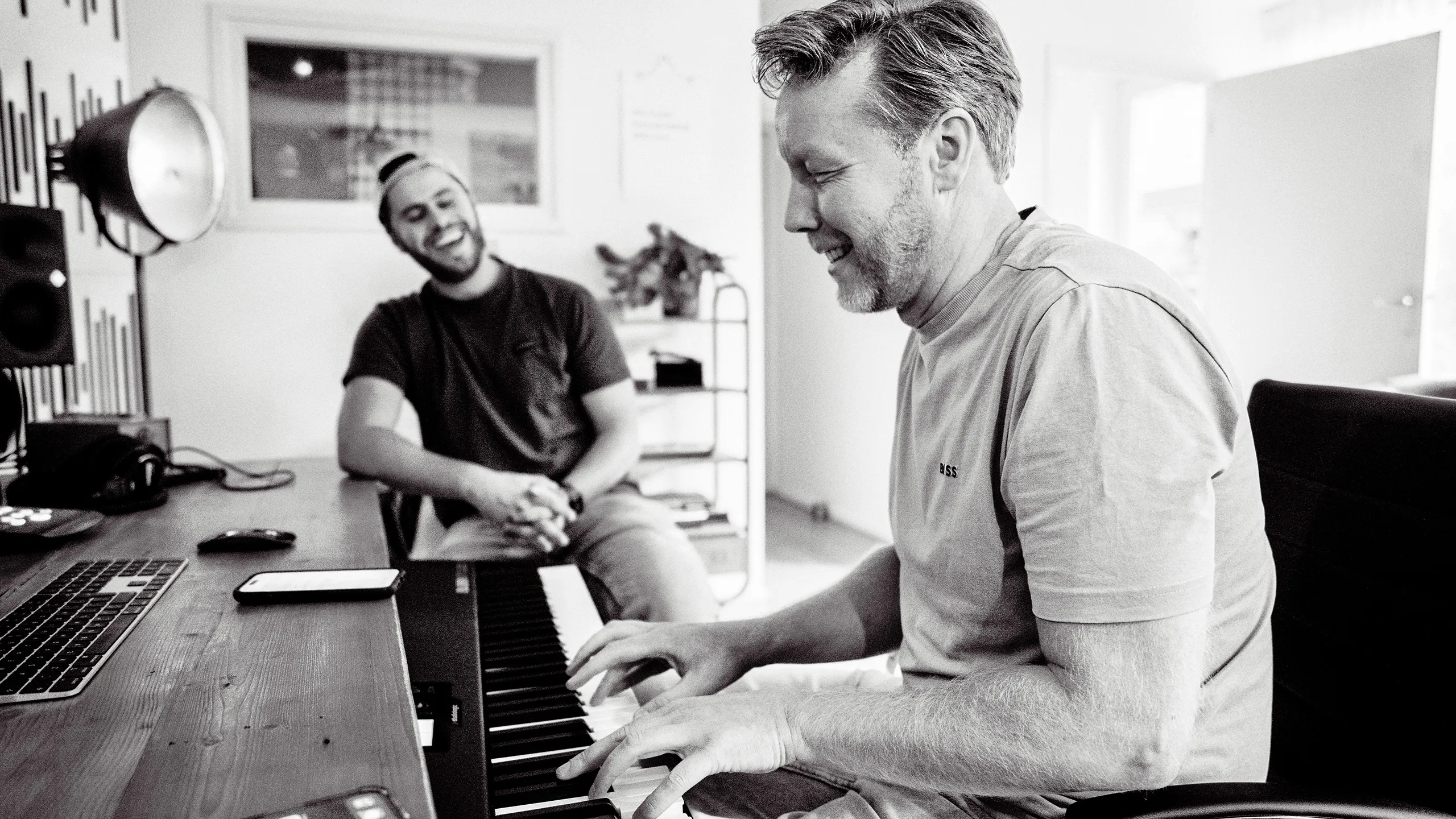 Adam Dekker en Artiest in een studio, waarbij Adam op de voorgrond piano speelt en de Artiest lachend toekijkt.