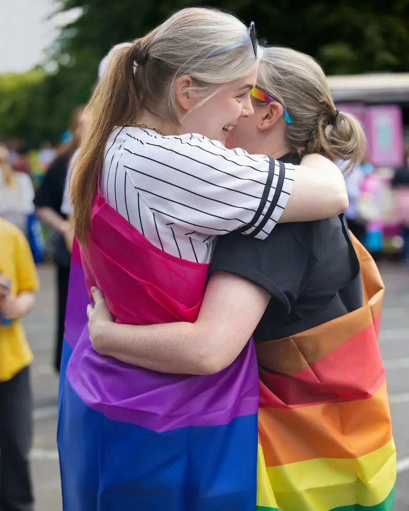 Two friends hugging at a Pride event, wrapped in bisexual and rainbow flags