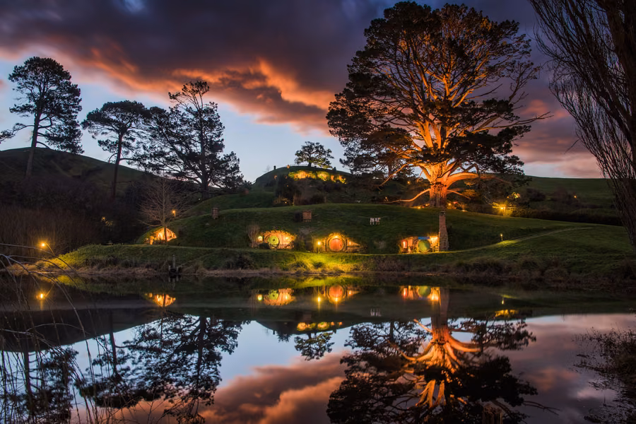 Hobbit holes with lit windows reflected in the lake at dusk at the Hobbiton Movie Set near Matamata