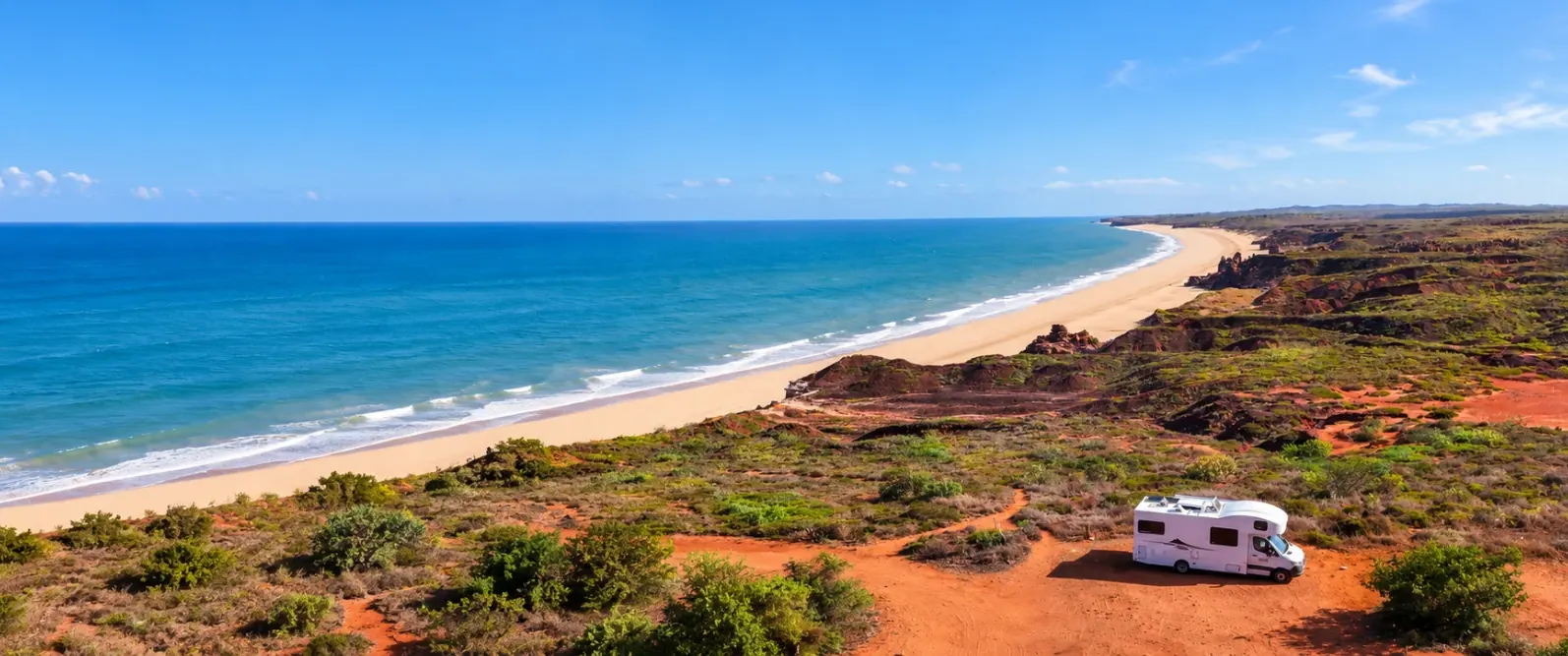 White motorhome parked on red pindan dirt on a clifftop above a long sandy beach and blue ocean in the Kimberley
