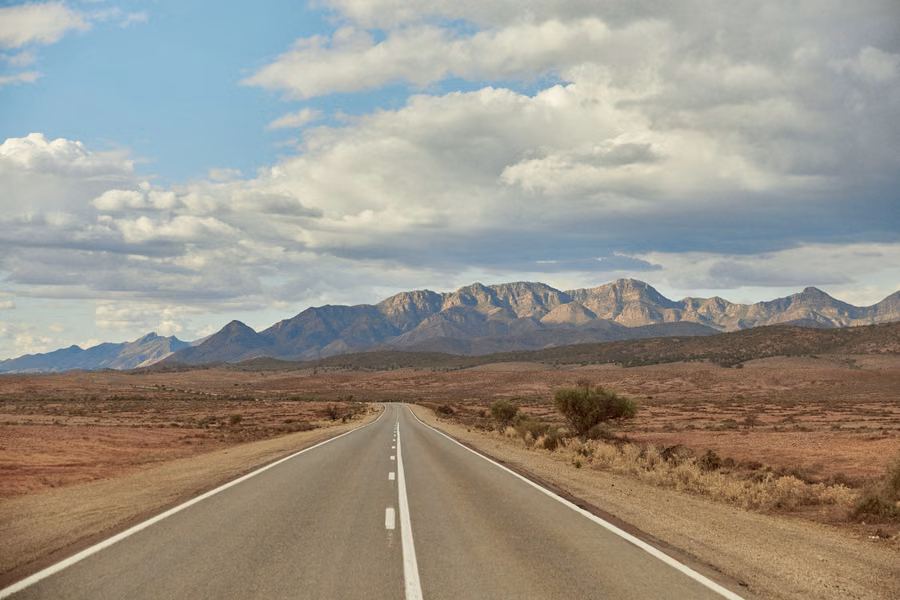 Straight sealed road leading across red plains to the rugged peaks of the Flinders Ranges under a cloudy sky