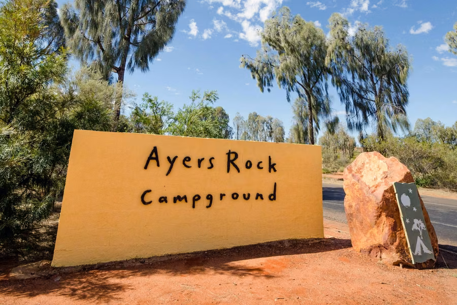 Ayers Rock Campground entrance sign beside a red boulder and desert oaks at Yulara
