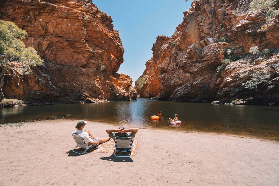 Visitors relaxing on the sandy shore of the Ormiston Gorge waterhole between towering red cliffs