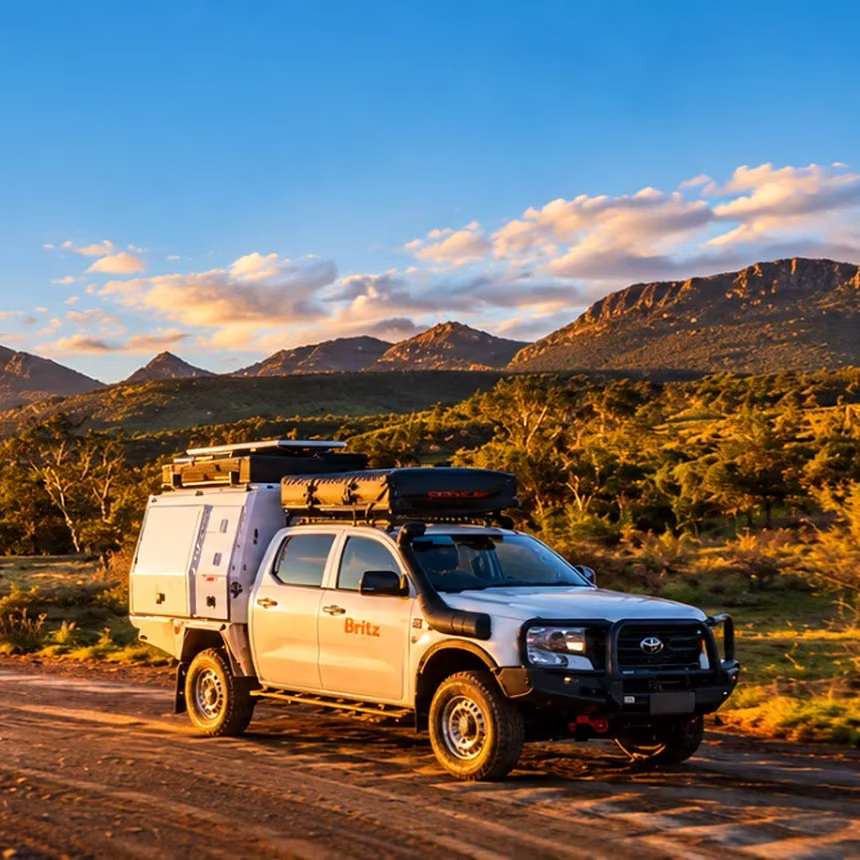 White Britz 4WD camper with a rooftop tent on a dirt road below rugged ranges at sunset