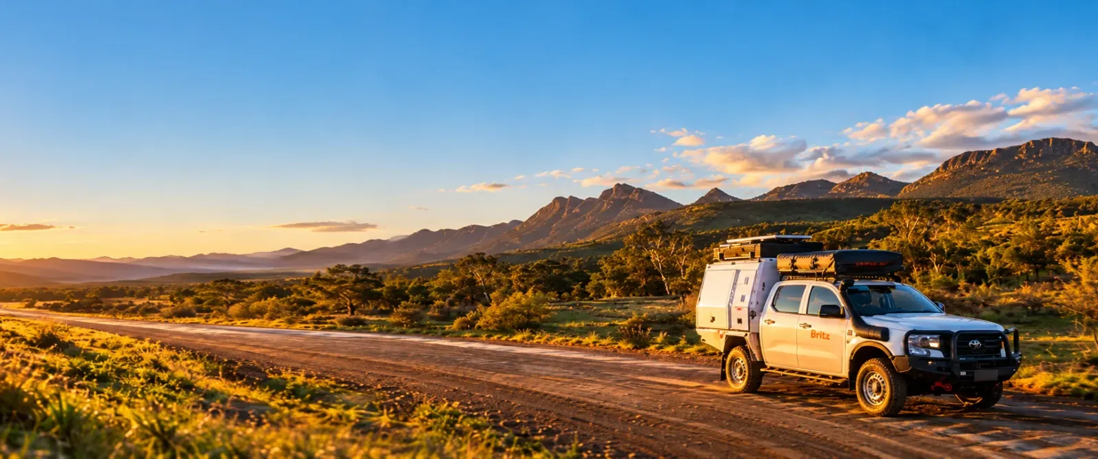 White Britz 4WD camper with a rooftop tent on a dirt road below rugged ranges at sunset