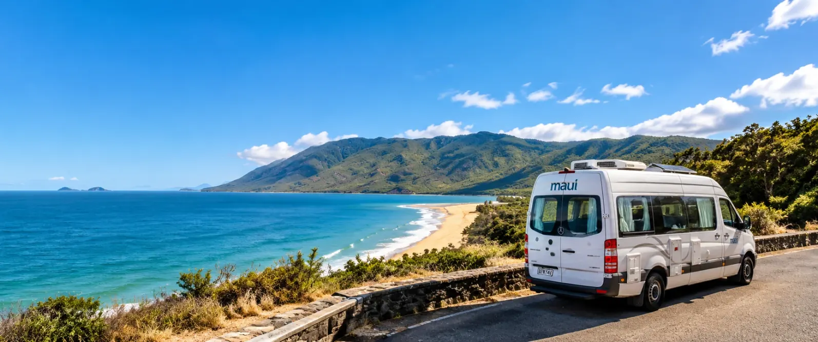 White maui campervan parked at a roadside lookout above a sandy beach, turquoise sea and green mountains