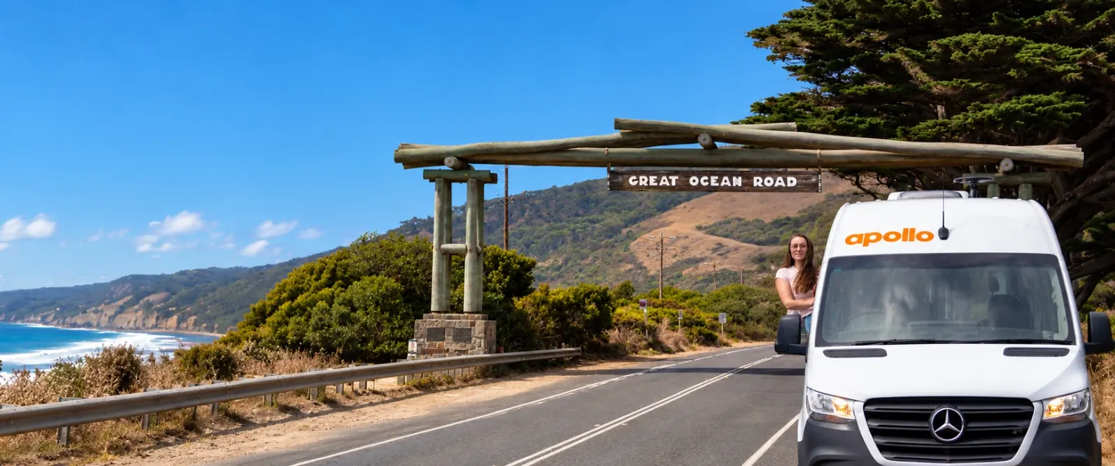 Woman leaning from the window of a white Apollo campervan on a coastal road beside the timber Great Ocean Road arch