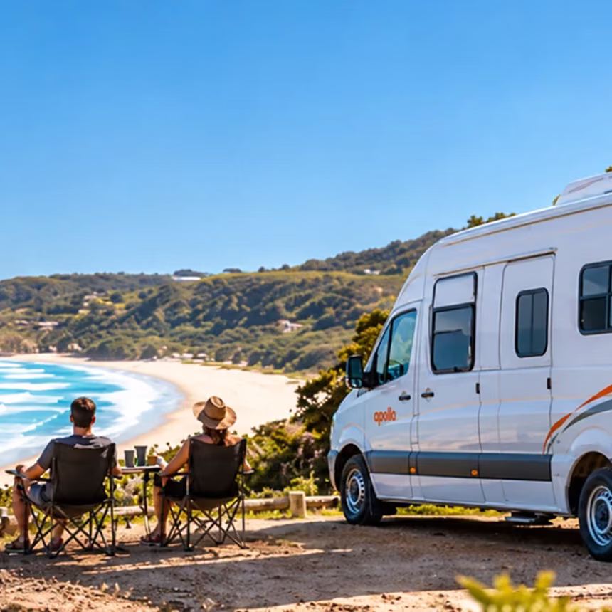 Couple in camp chairs beside an Apollo campervan on a bushy clifftop above a curving surf beach