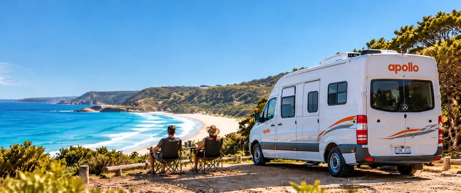 Couple in camp chairs beside an Apollo campervan on a bushy clifftop above a curving surf beach