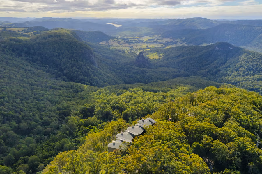 Aerial view over rainforest at Binna Burra, with a row of lodge roofs on the ridge and the valley and Egg Rock beyond
