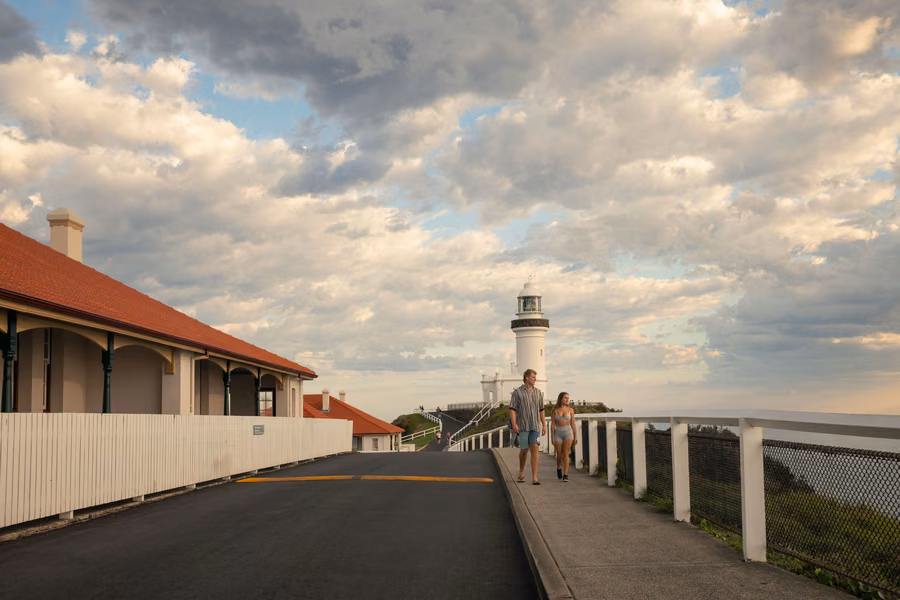 Two people walking up the path beside the keepers' cottages towards Cape Byron Lighthouse under a cloudy sky