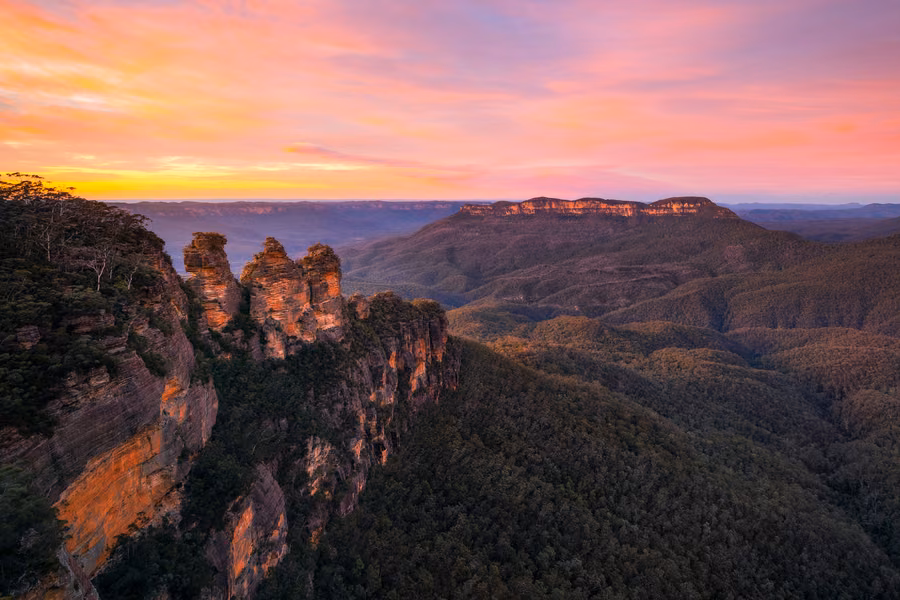 The Three Sisters rock formation above the forested Jamison Valley under a pink and orange sunrise sky in the Blue Mountains