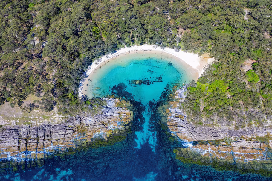 Aerial view of the turquoise, almost round Honeymoon Bay ringed by white sand, rock shelves and coastal bushland at Jervis Bay