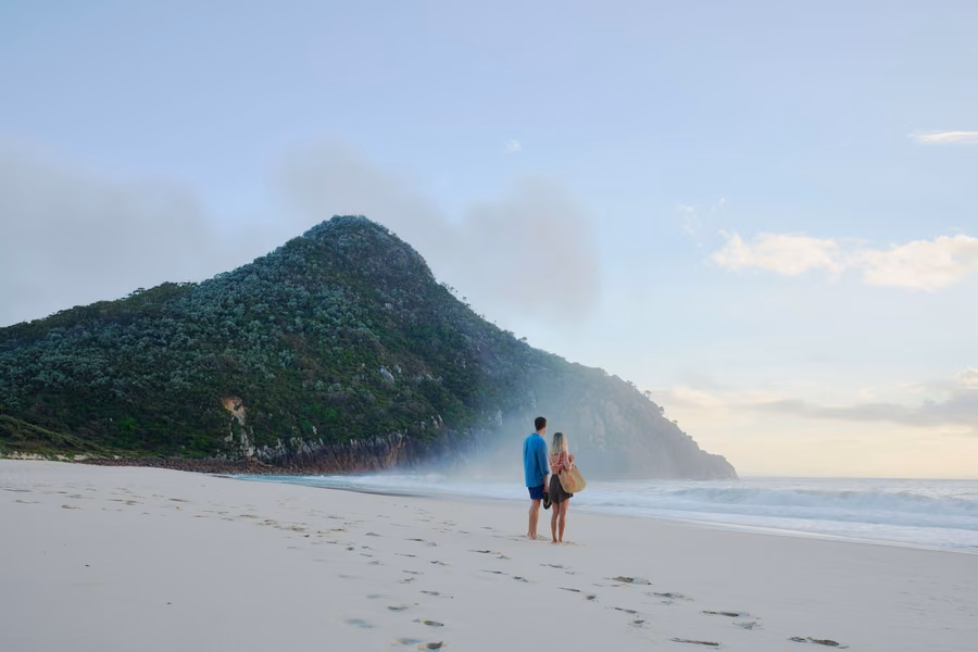 Couple standing on Zenith Beach looking towards a steep bush-covered headland and sea mist in Port Stephens