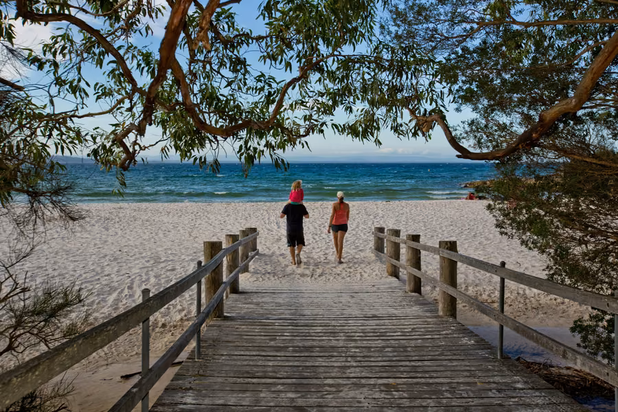 Family walking from a timber boardwalk onto the white sand at Green Patch beach in Booderee National Park, framed by trees