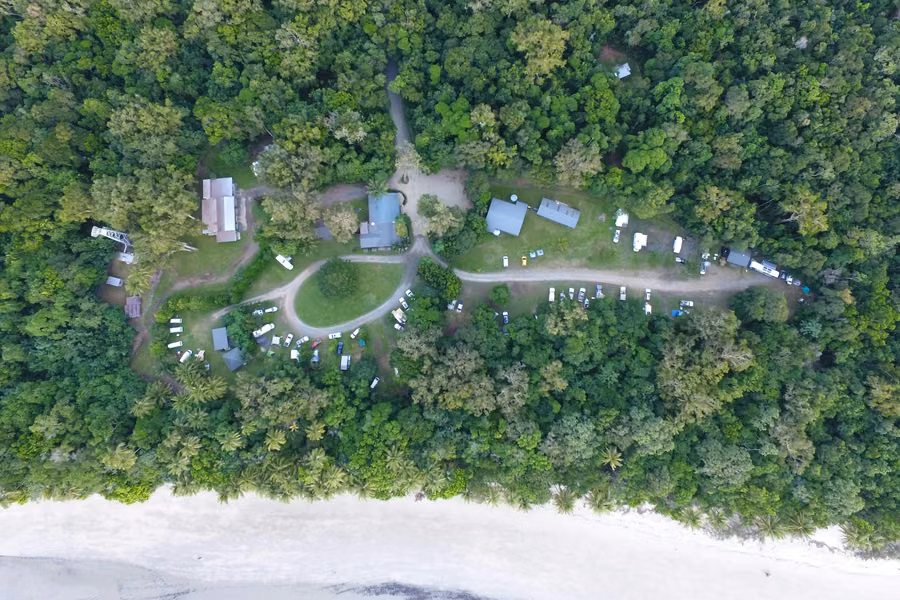 Top-down aerial view of Cape Tribulation Camping, with campsites in a rainforest clearing just behind a white sand beach