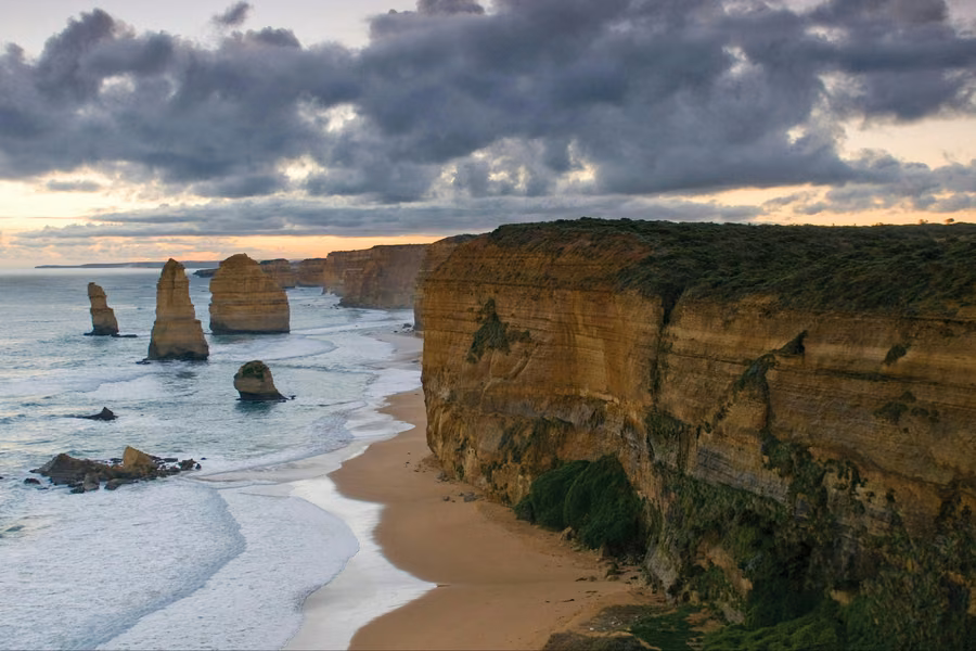 Limestone stacks of the Twelve Apostles beside tall cliffs and a sandy beach under a cloudy sunset sky