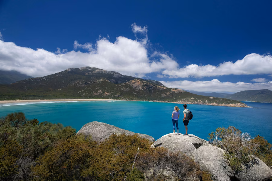 Two hikers on granite boulders above a turquoise bay and mountains at Wilsons Promontory