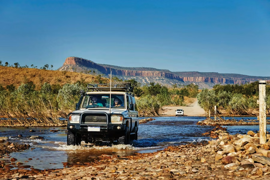 4WD with a roof rack driving through the shallow, rocky Pentecost River crossing on the Gibb River Road, with the Cockburn Range behind