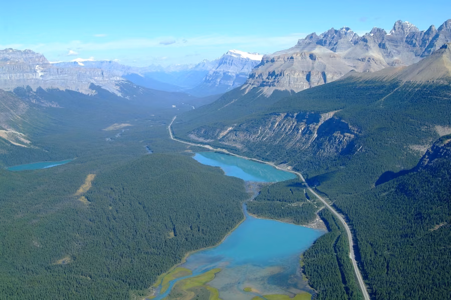 Aerial view of the Icefields Parkway running along a forested valley past turquoise lakes below jagged peaks