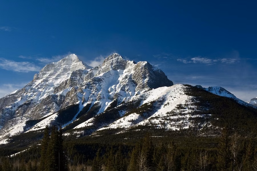 Snow-streaked mountain peak rising above dark forest under a deep blue sky in Kananaskis Country