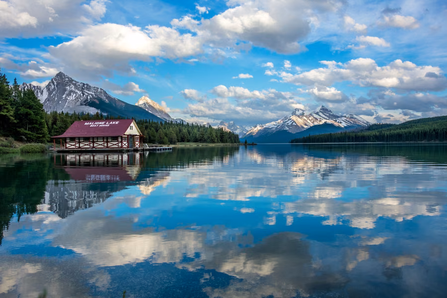 Maligne Lake boathouse and snow-capped peaks reflected in calm blue water under scattered clouds in Jasper National Park