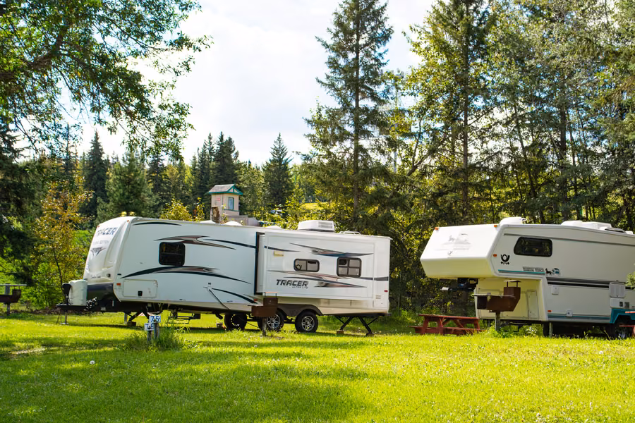 Two travel trailers parked on a grassy site among tall spruce and poplar trees at Rainbow Valley Campground