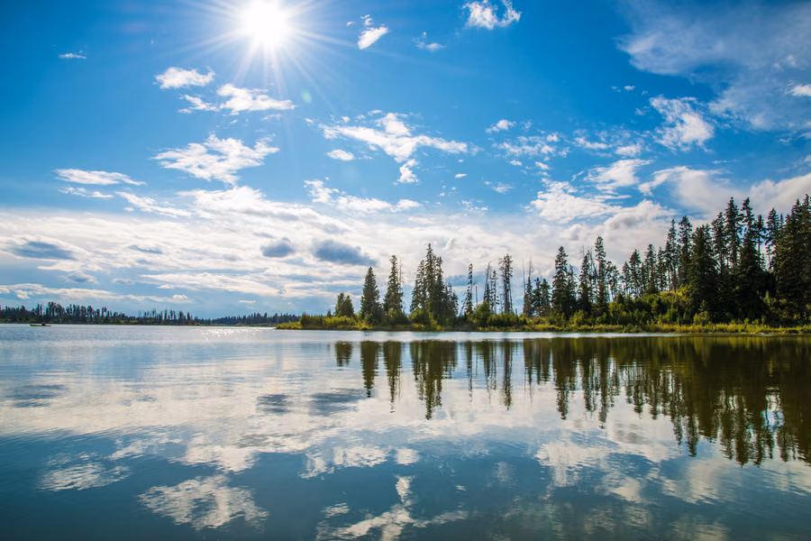 Calm lake reflecting spruce trees and white clouds under a sunny blue sky in Elk Island National Park