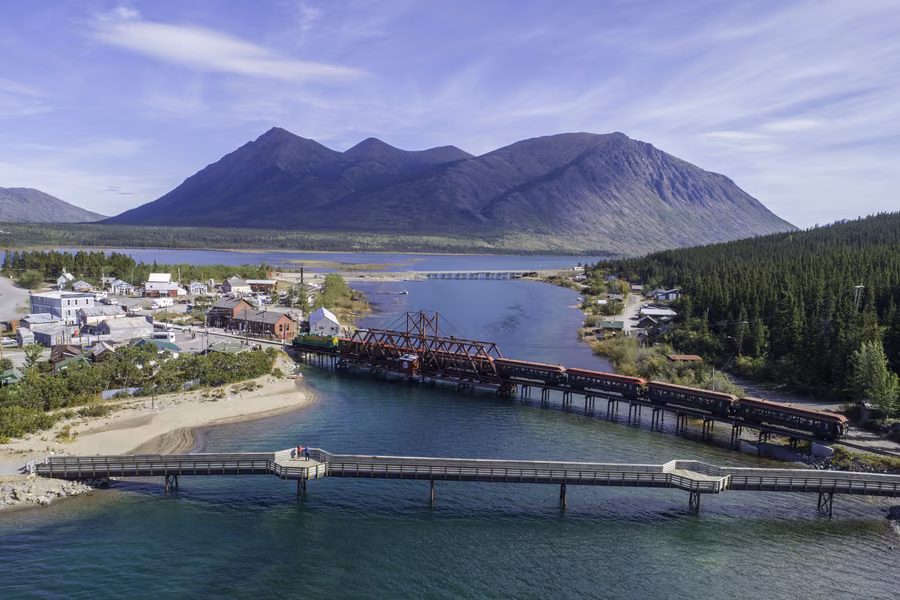 Aerial view of Carcross, with a train on the red rail bridge between two lakes and mountains behind