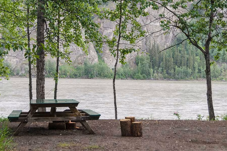 Picnic table and log seats under leafy trees on the bank of the Yukon River, with cliffs on the far side